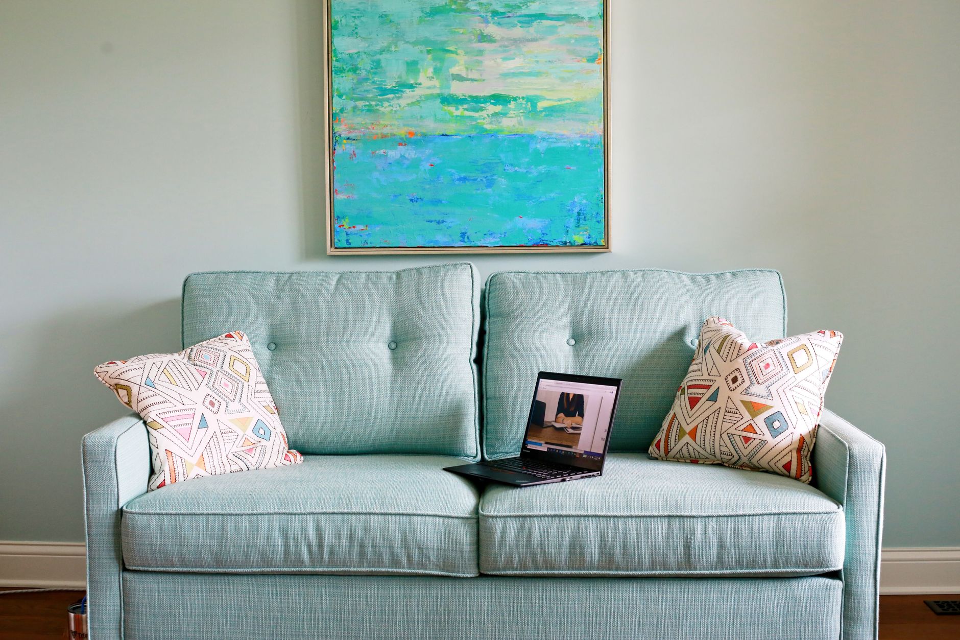 toile abstraite aux tons turquoise et vert d'eau accrochée au-dessus d'un canapé en tissu bleu-vert avec deux coussins à motifs