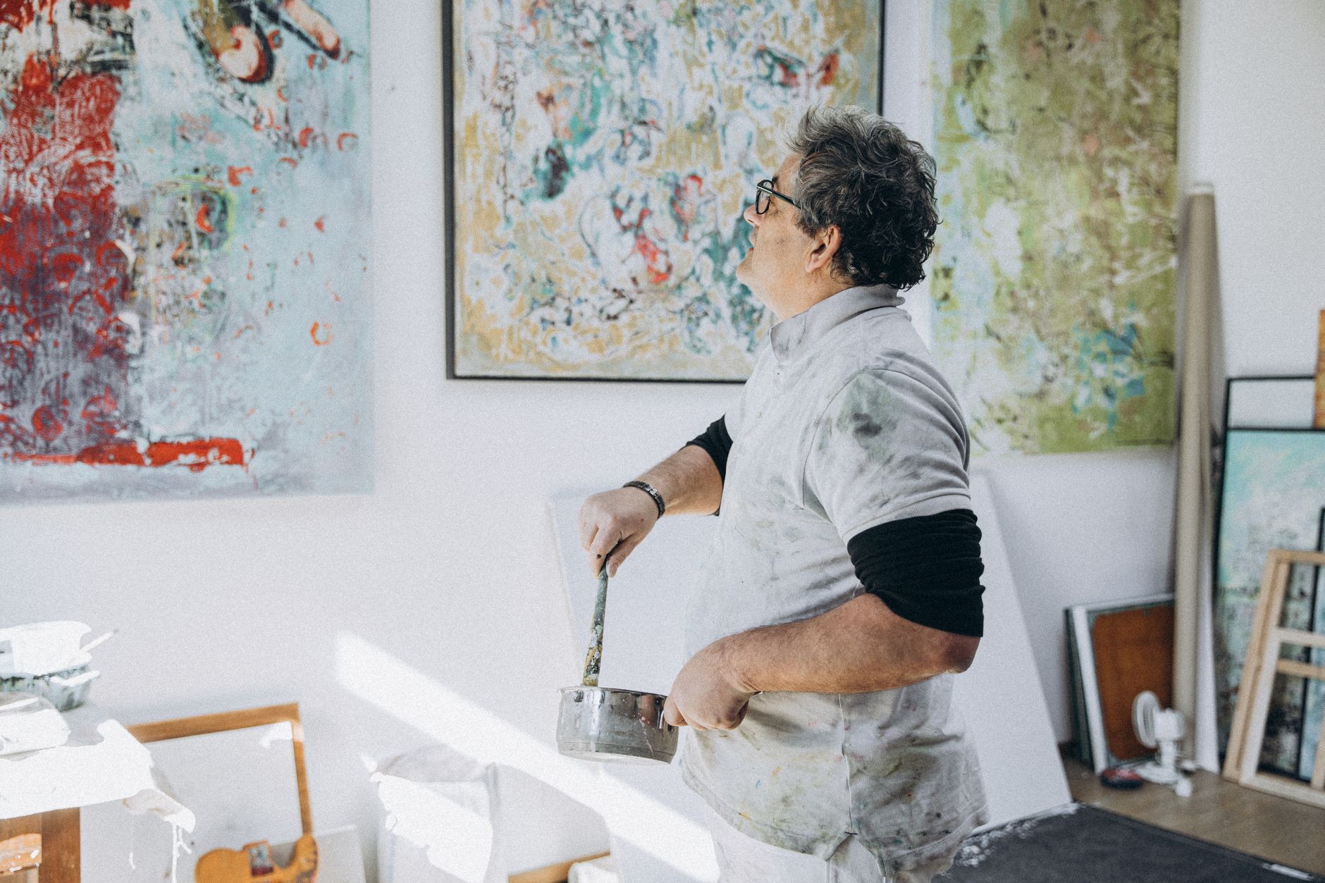 peintre en tenue d'atelier tachée de peinture, un pot à la main, observant ses grandes toiles abstraites accrochées au mur